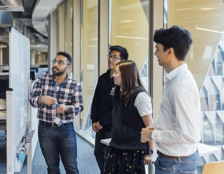 Three people watching as a fourth person demonstrates something on a white board