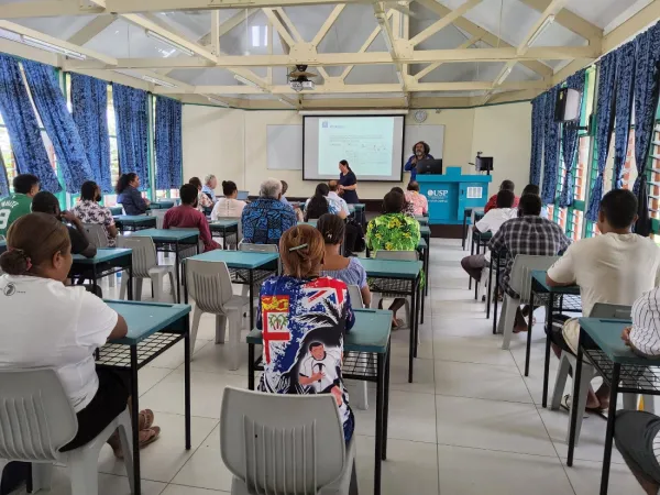 People seated at desks listening to a presentation from the front of the room.