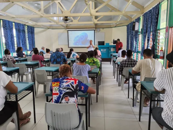 People seated at desks listening to a presentation from the front of the room.